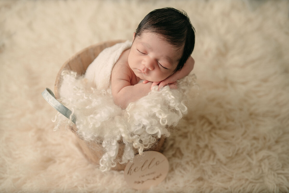 Newborn baby with thick brown hair laying in bucket resting cheek on hands