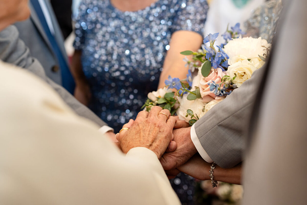 A close-up of hands being held during a wedding ceremony, showcasing the bride's bouquet and the hands of their loved ones.