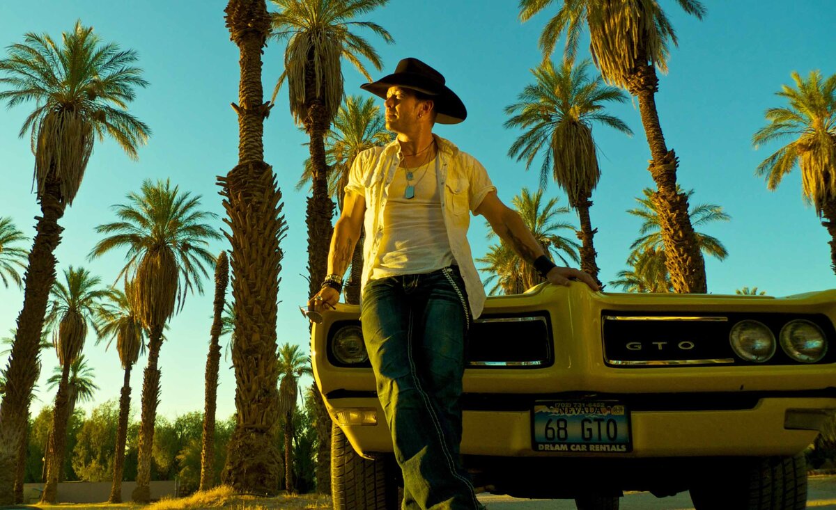 Male musician portrait Aaron Pritchett wearing jeans white shirt black cowboy hat leaning against yellow convertible front grill palm tress blue sky background