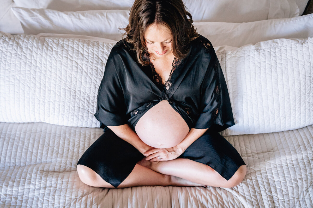 A pregnant woman sits cross-legged on a bed, gazing down at her belly, her hands resting gently on it, wrapped in a black robe.