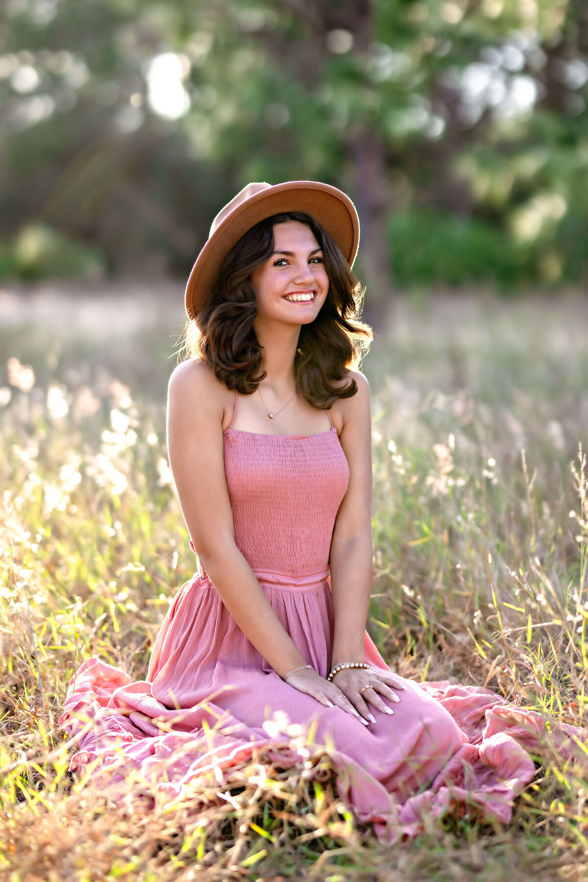 Adorable senior girl in long pink dress sitting with hands in lap softly giggling at the camera during sunset in Central Florida