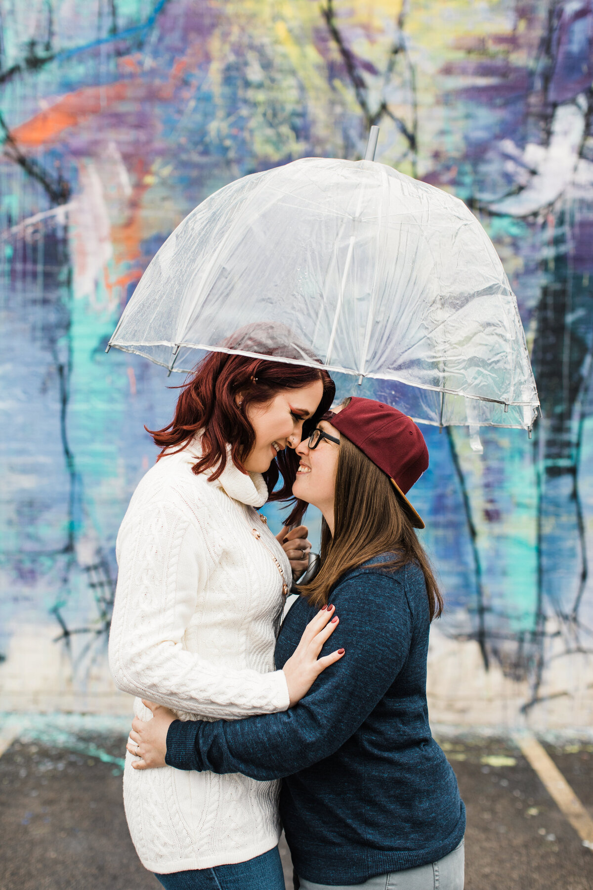A lesbian couple holding each other close under an umbrella and in front of a colorful mural during their engagement session in Deep Ellum in Dallas, Texas. The woman on the right is wearing a dark blue sweater, glasses, and a backwards red baseball cap. The woman on the right is wearing a detailed white sweater and jeans.