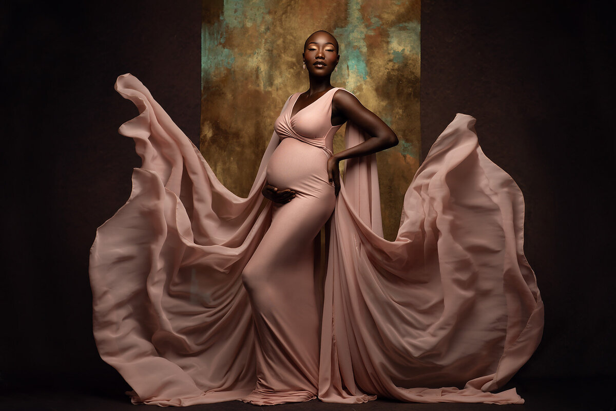 A captivating maternity photograph featuring an expecting mother in a flowing blush pink gown with billowing fabric. The gold and teal textured backdrop enhances the luxurious feel of this DFW maternity photography session.