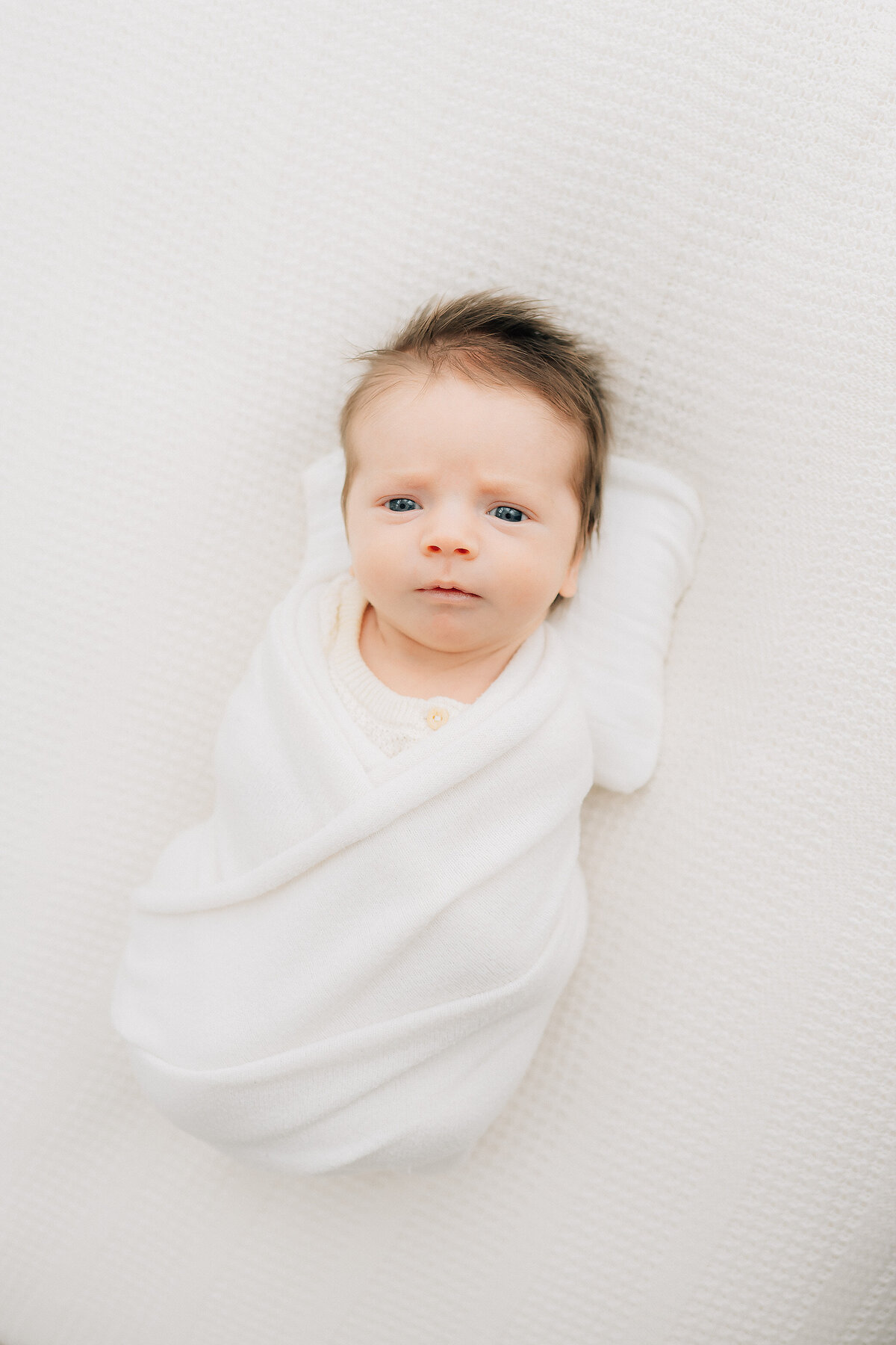 swaddled baby in white with full hair looking up at photographer