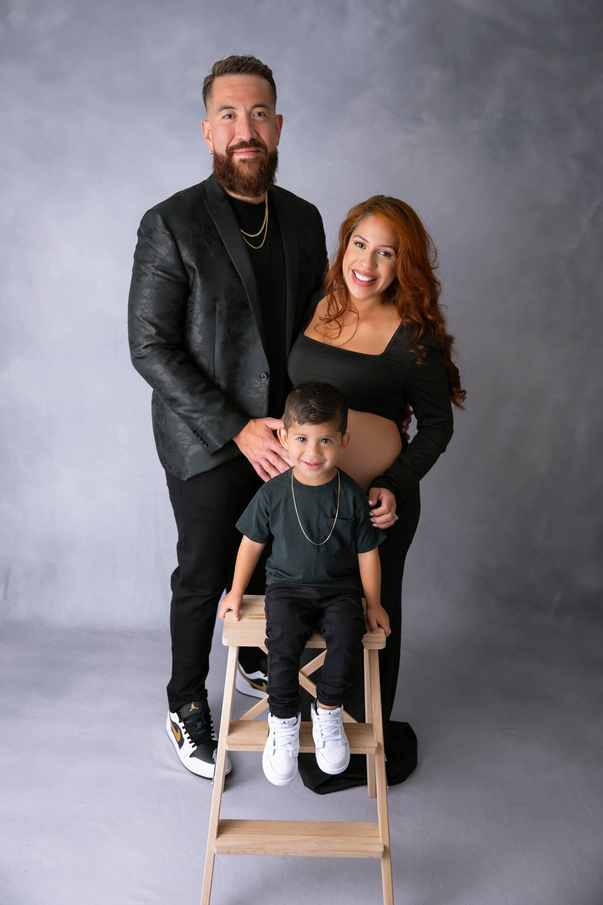 Expecting parents pose with their smiling young son sitting on a step stool, all dressed in black.