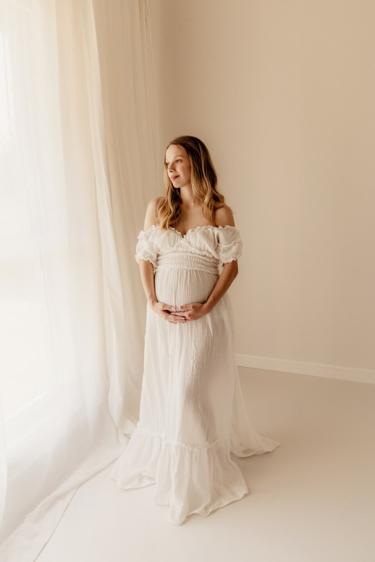 Pregnant woman with blonde hair wearing a long white dress looking out a window in Yukon, Oklahoma.