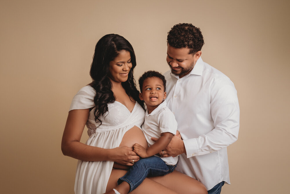 Maternity session with Atlanta, Georgia maternity photographer pregnant woman holding baby bump wearing white sheer maternity  dress on white backdrop posing with husband and 2 year old son  on cream color backdrop