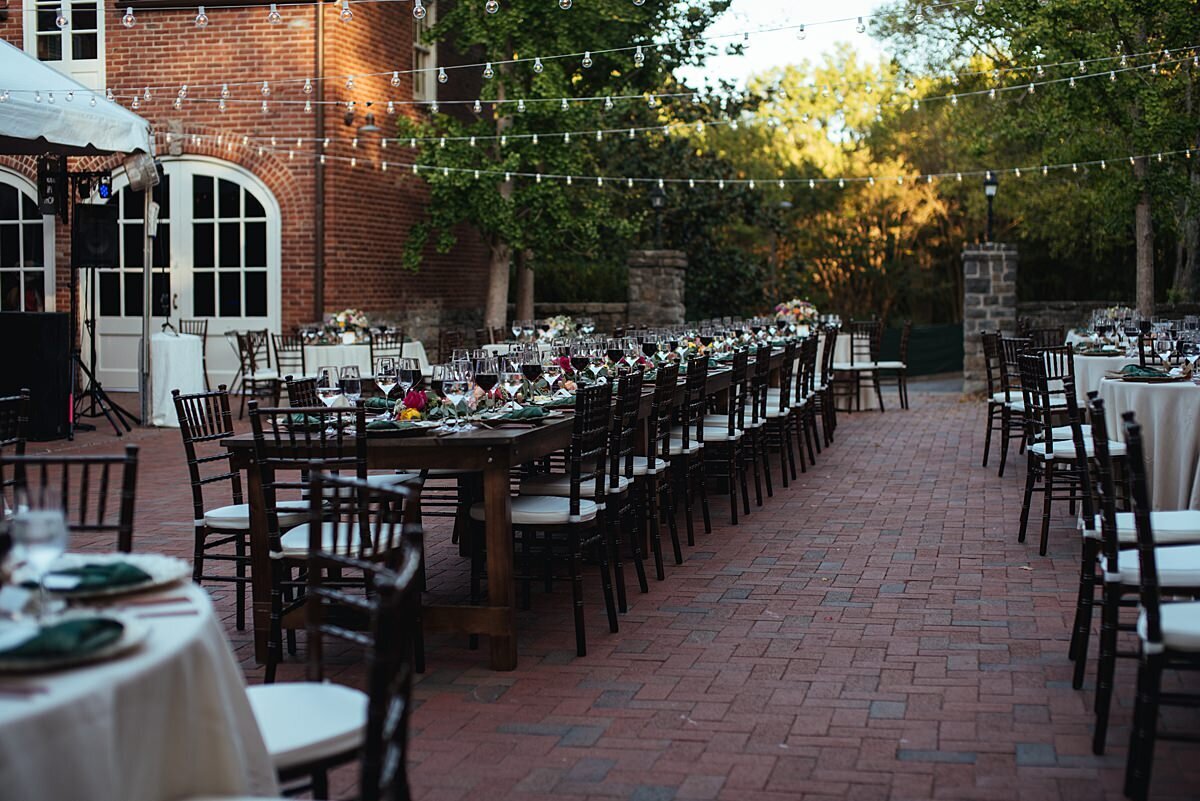 A wedding reception at the Frist Learning Center courtyard at Cheekwood Botanical Gardens. The assortment of round and long tables have ivory table cloths and brown chiavari chairs in an exposed brick courtyard with hanging string lights above.