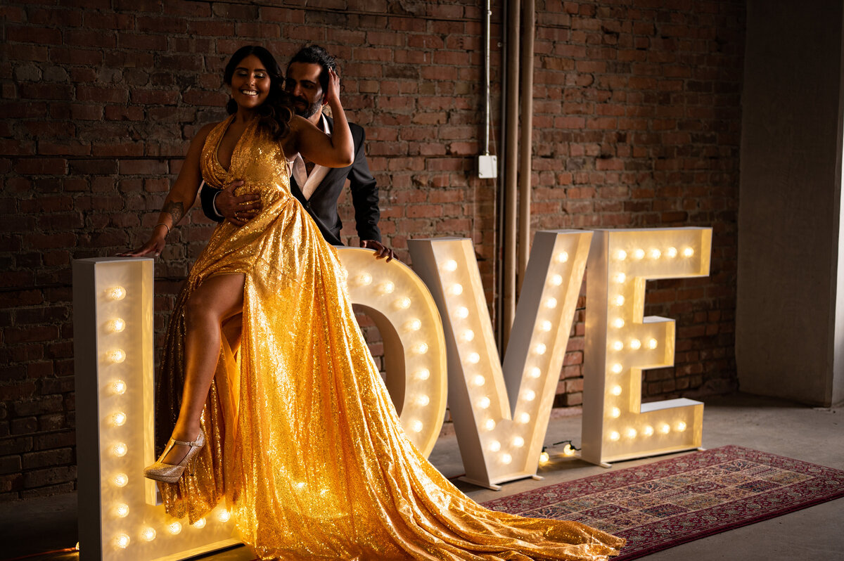 Couple sits on a LOVE sign in a rustic buidling for the engagement session in Edmonton
