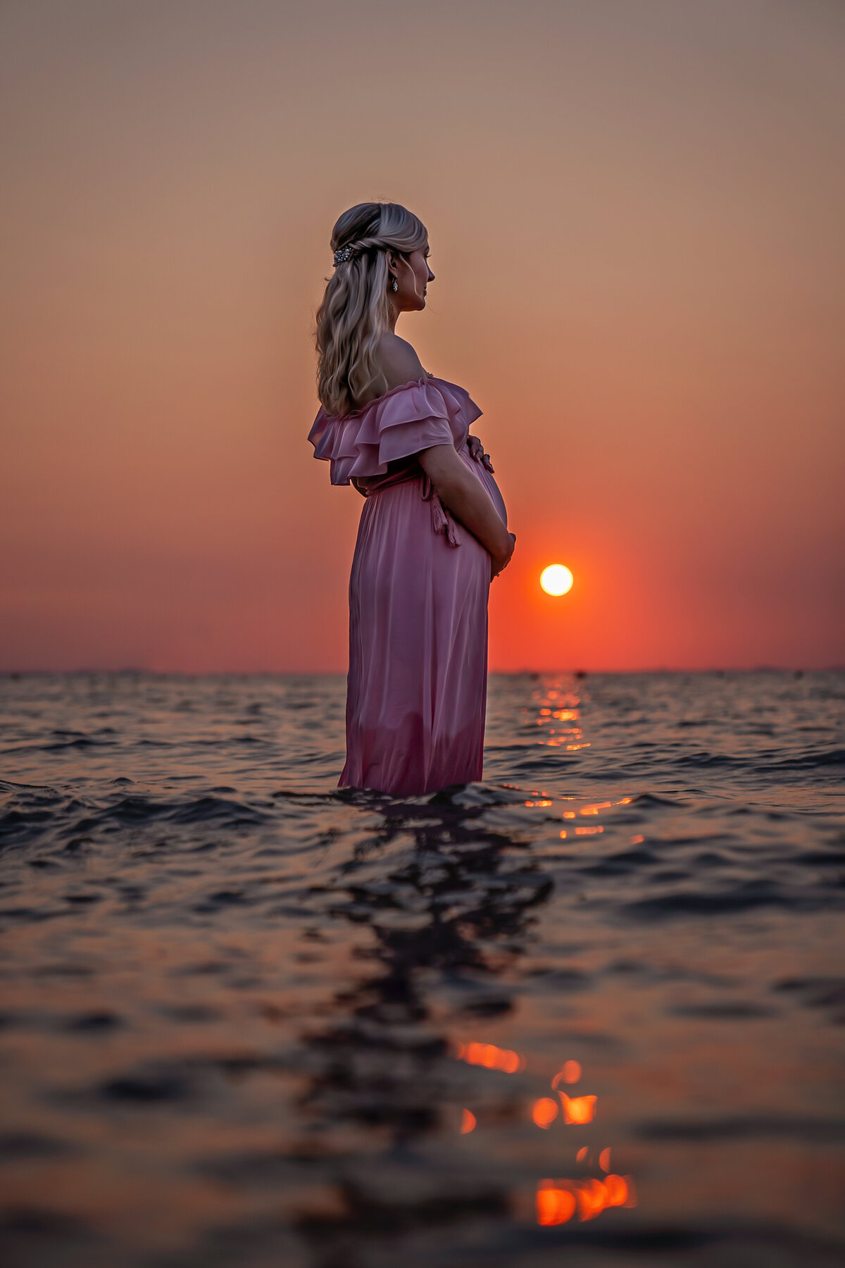 gorgeous pink sunset and pregnant mom in pink long maxi dress  in lake michigan waters  looking in to the distance.