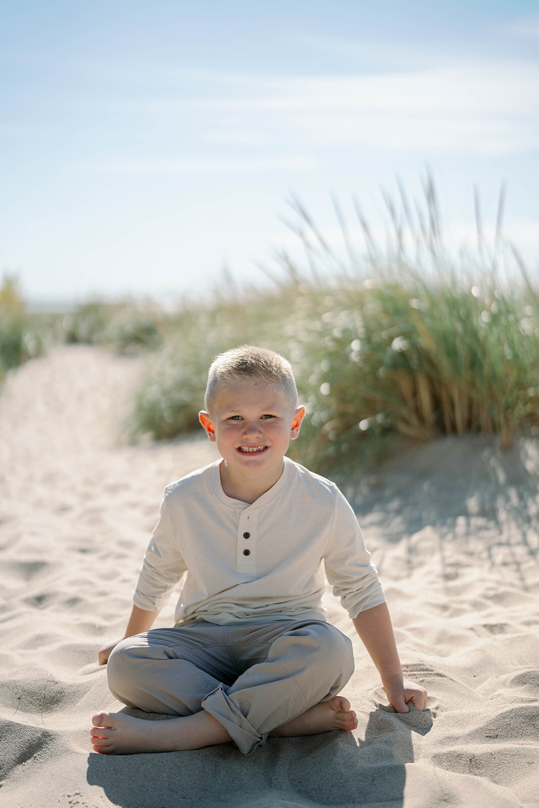 A young boy sitting cross-legged in the sand at Weko Beach smiling during the family’s mini session in Bridgman Michigan.