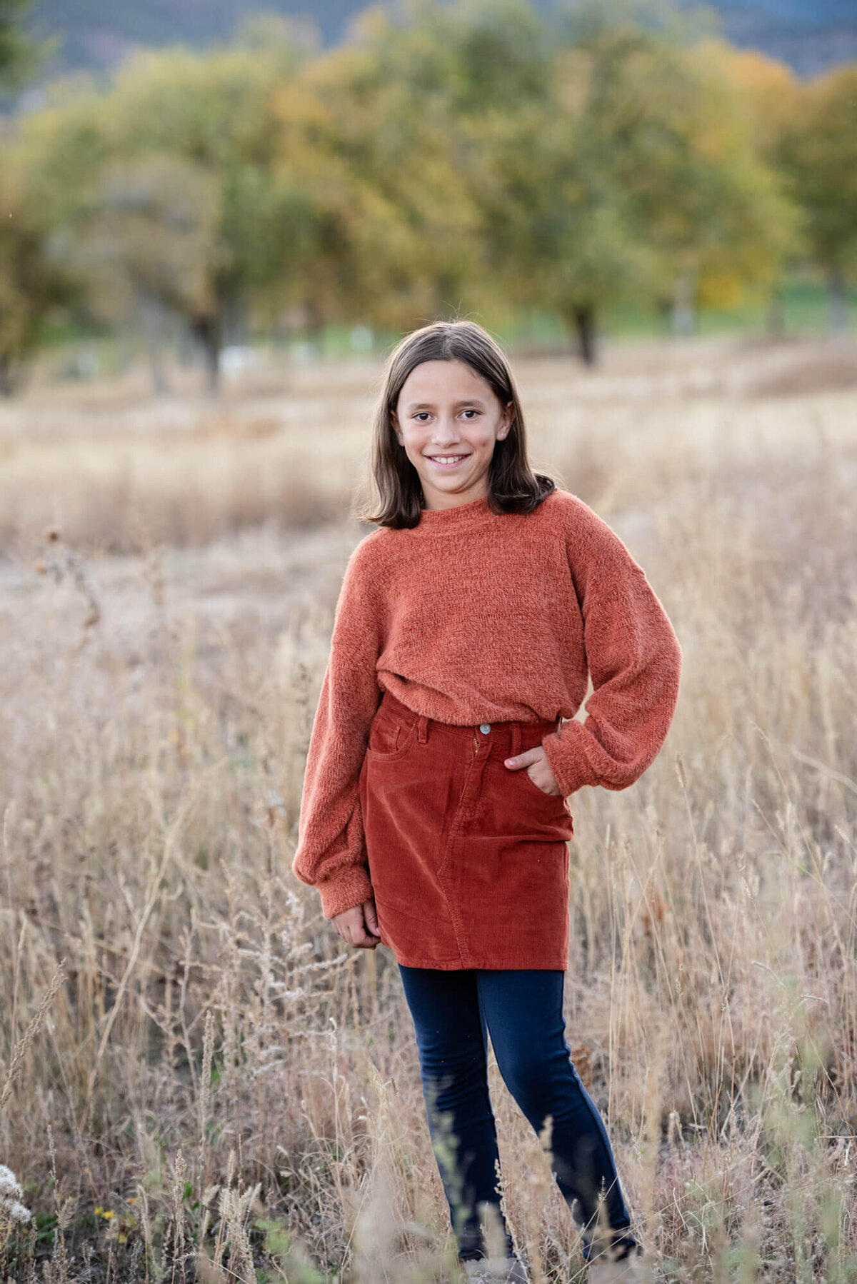 A young girl with dark hair in a red skirt stands in a field with a hand in her pocket