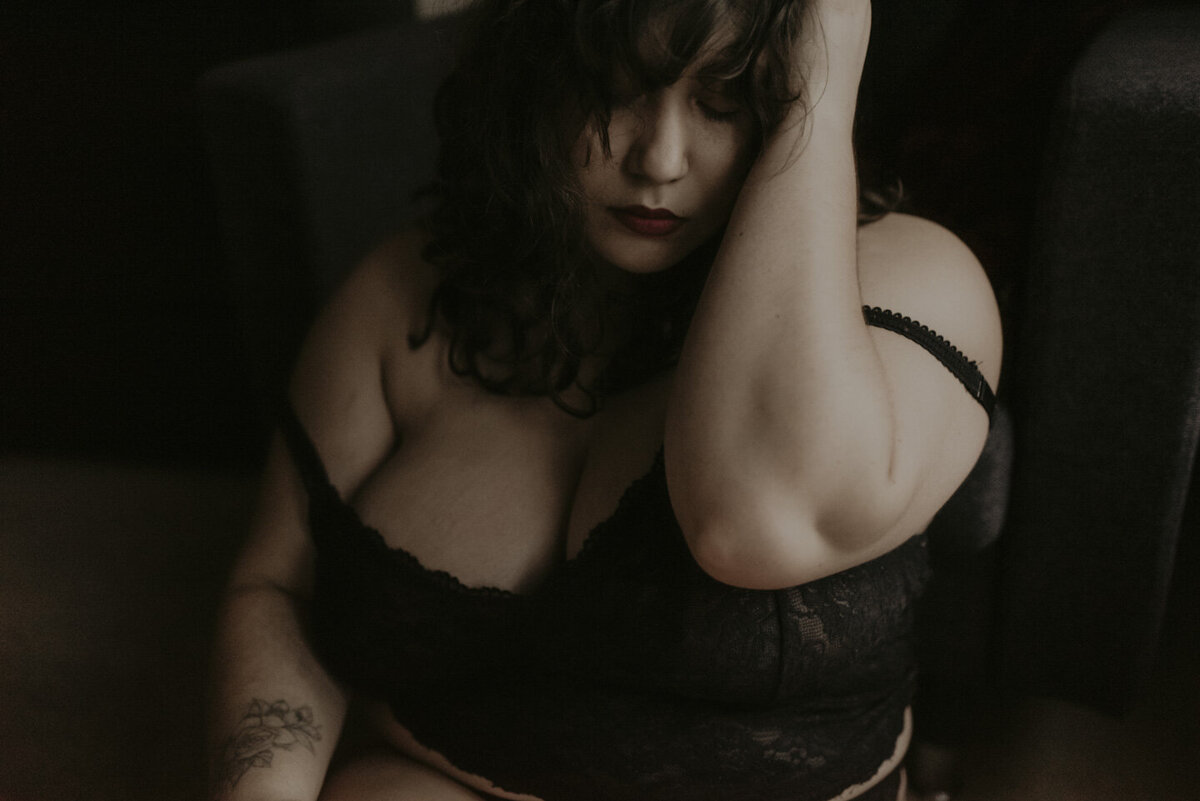 taeya sitting on the ground in front of a chair. She is in lingerie and her bra straps are hanging off the shoulders. Her left hand is up in her hair. Window light is coming from the right side of the frame.