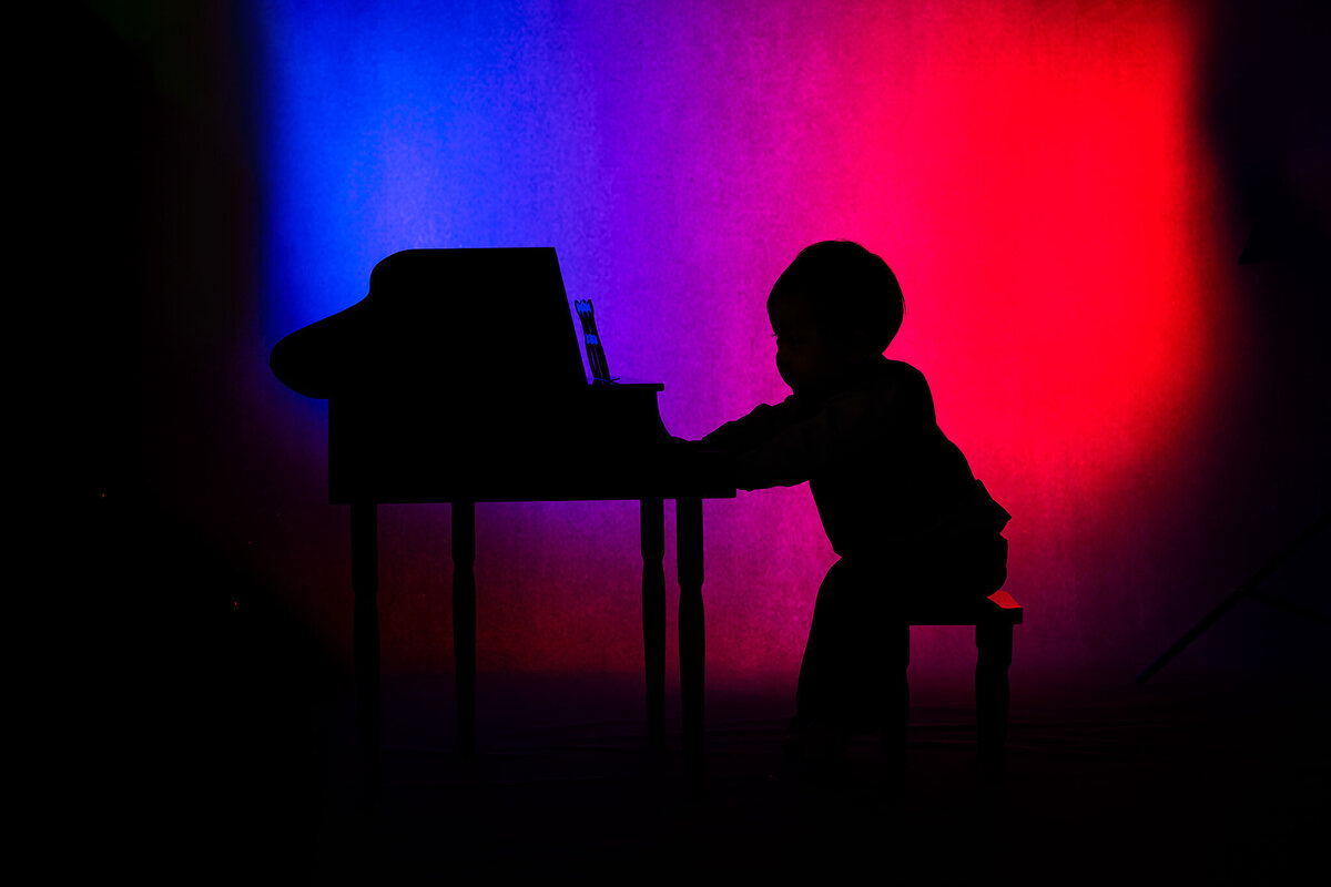 Dallas baby milestone photographer captures baby silhouetted at piano with colorful lighting.