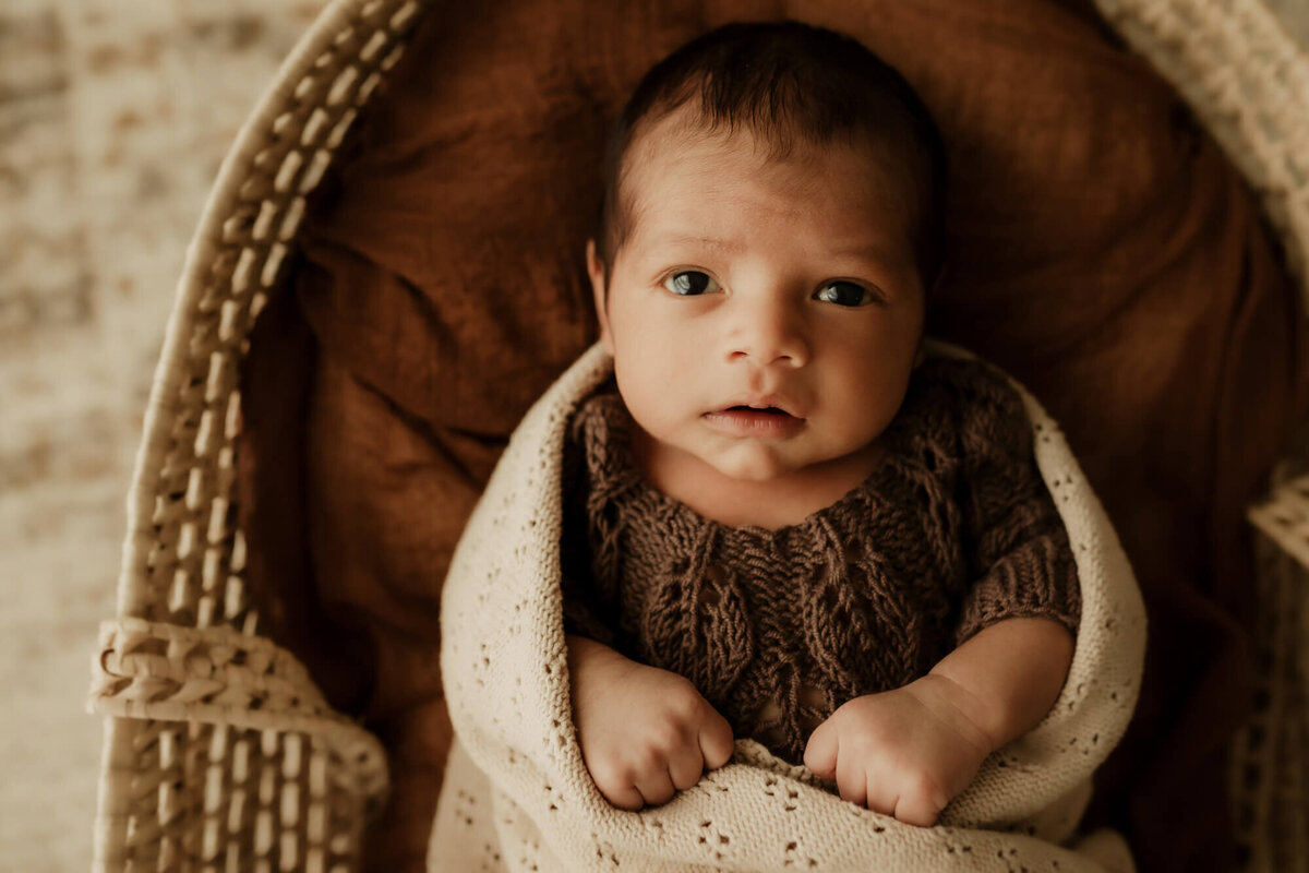 Baby boy wearing a brown knit romper in a a basket wrapped in a blanket in Oklahoma City.