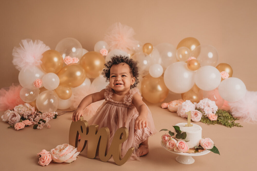 Cake smash session with Atlanta, Georgia cake smash photographer of one year old baby girl in pink tulle dress on tan background with neutral colored balloon garland eating her white iced birthday cake with pink roses on it