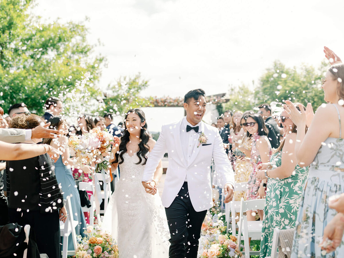 A joyful bride and groom walk down an outdoor aisle surrounded by guests throwing confetti. The couple smiles brightly under a clear sky, exuding happiness.
