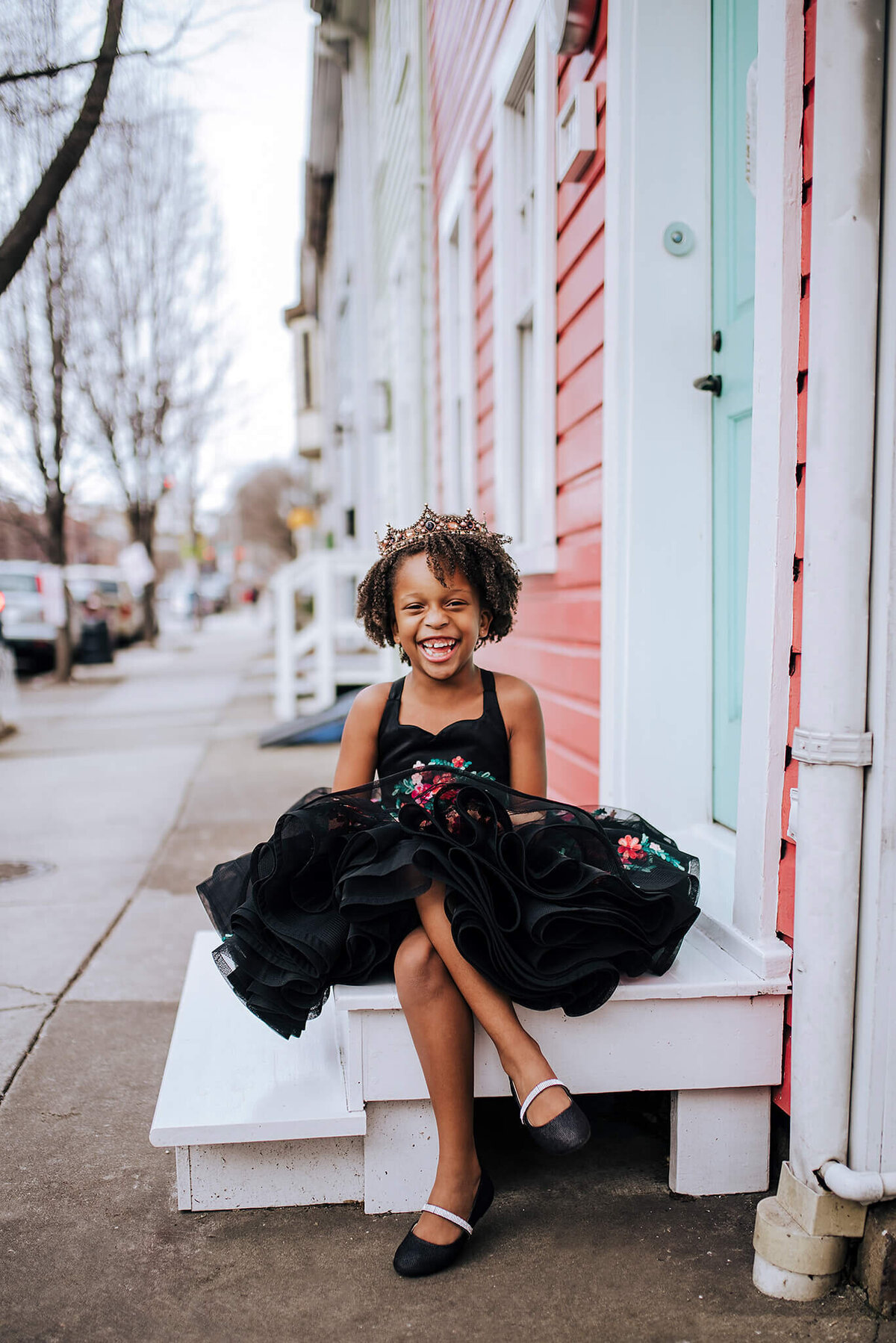 African American girl wearing a black and floral dress with black shoes sitting on a white step with a crown in Fells Point Baltimore Maryland