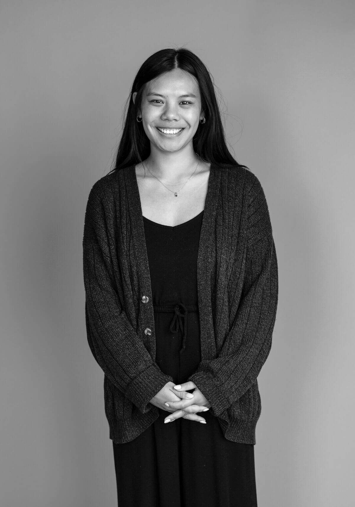 A black and white portrait of a woman in a cardigan, smiling gently with hands folded—capturing a natural moment before her transformation.