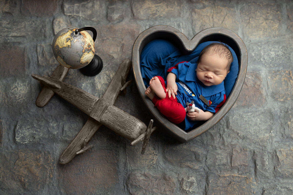 Newborn baby dressed in Southwest Airlines outfit, sleeping in a heart-shaped bowl next to a globe and wooden airplane – Dallas newborn photographer