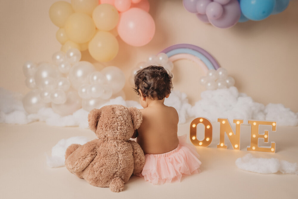 Photo shoot at Atlanta, Georgia cake smash photographer studio with one year old little girl in pink tutu smashing a cake with rainbow pastel balloon garland behind her with white fluffy clouds.