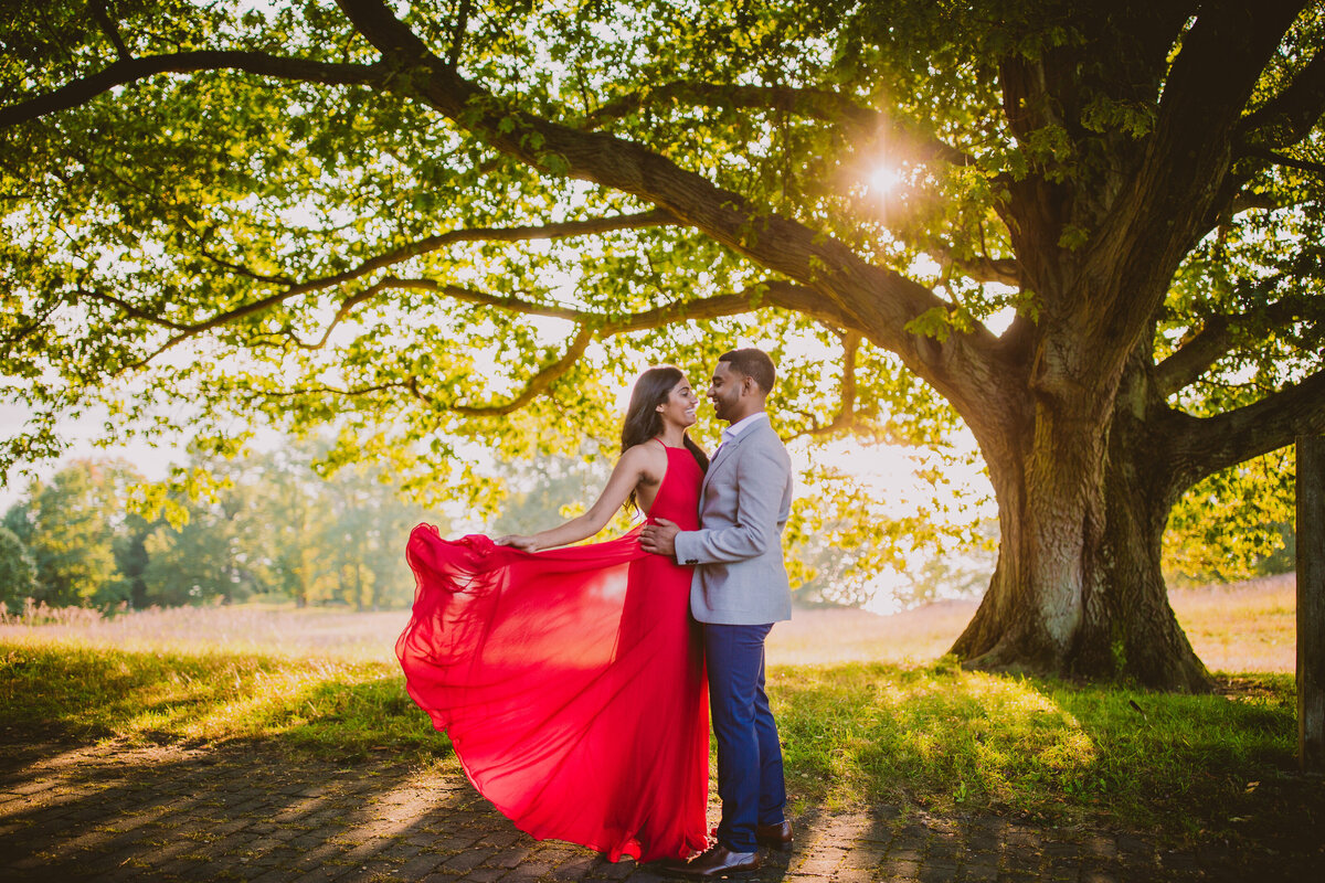 An engaged couple with red outfit flying in the wind near a tree at rockwood hall in pleasantville ny