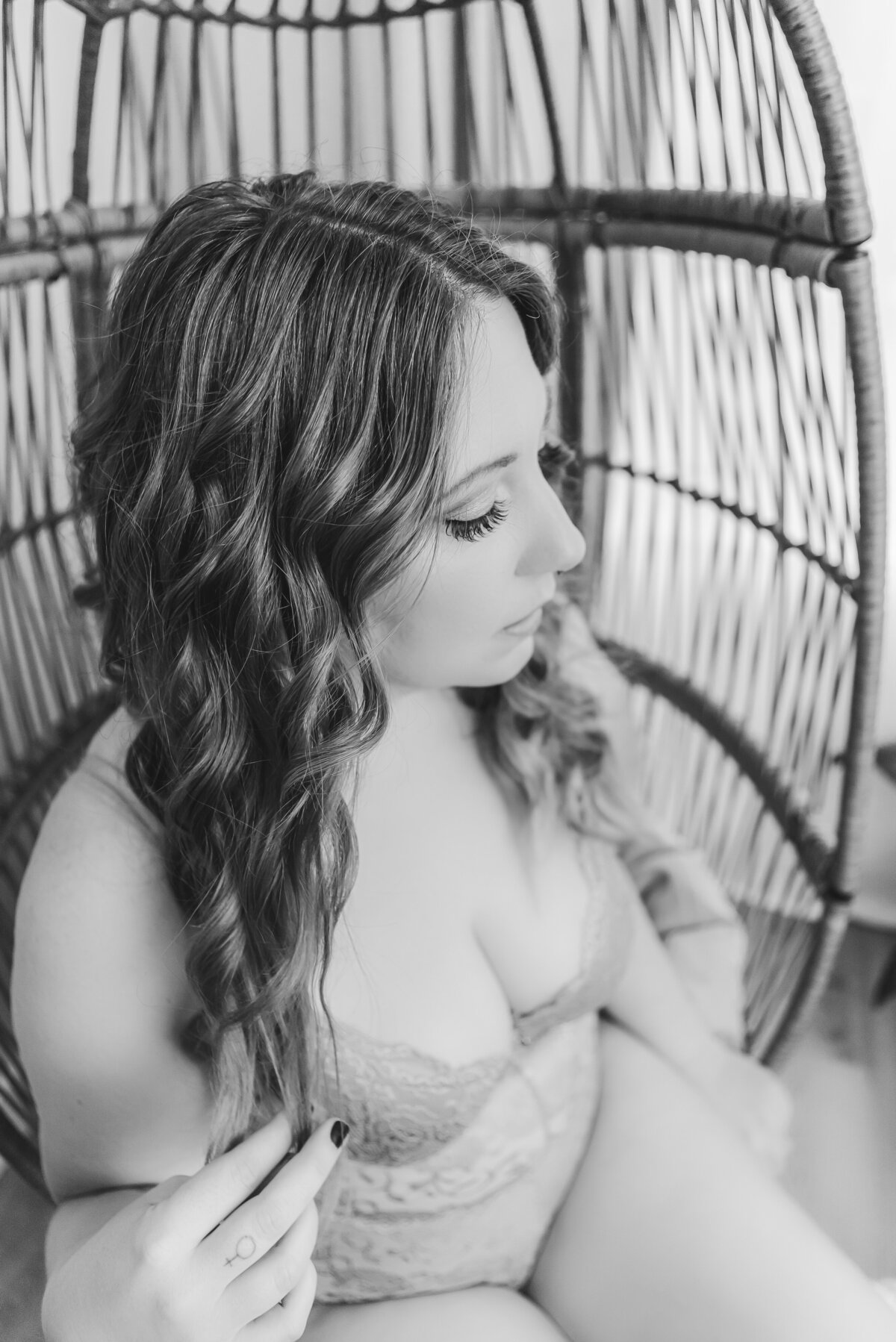 A woman sitting in a wicker chair, gently playing with her curled hair, wearing light gray lingerie.