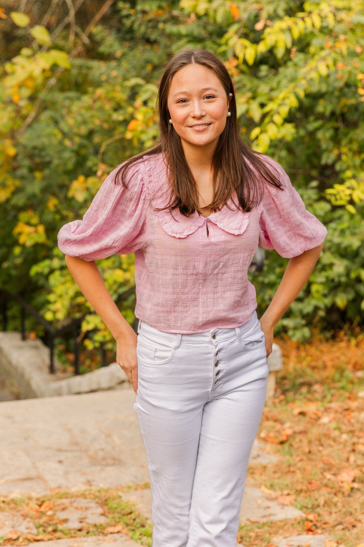 senior girl wearing white jeans and pink top stand ankles crossed and hands on back pockets on park path