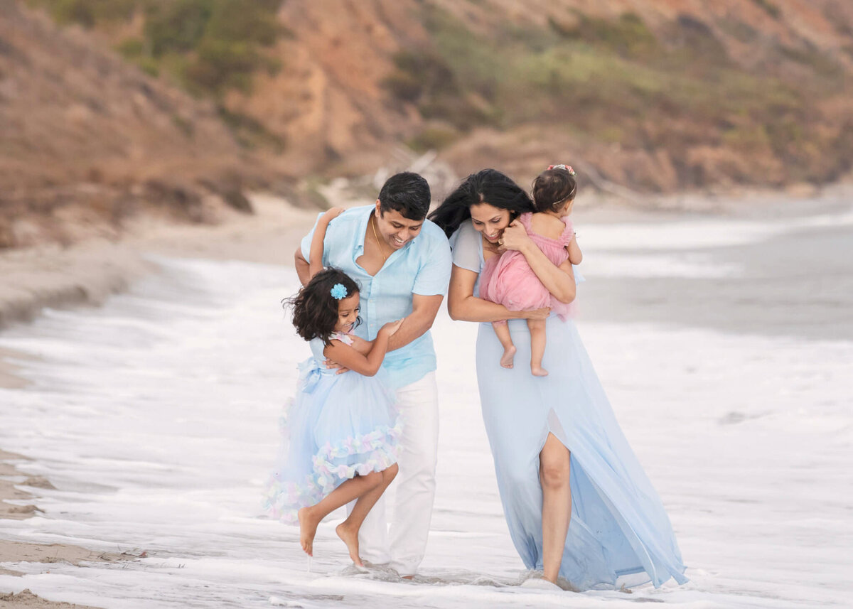 Mom, dad and two daughters laughing as the water covers their feet at the beach in Mallibu photographer by Los Angeles Photography Elsie Rose