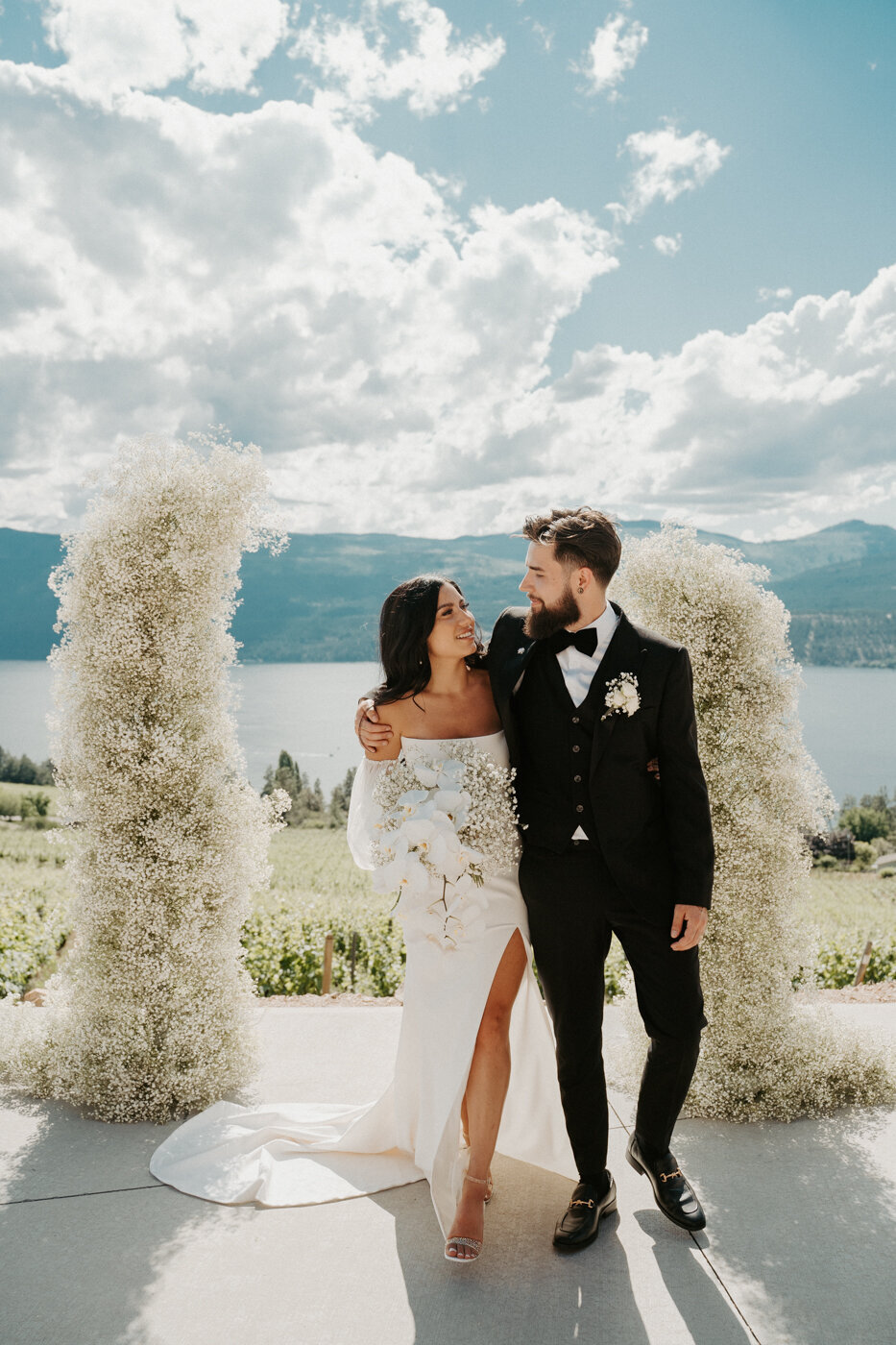 Kelowna Wedding Couple over looking the Okanagan Lake in Lake Country BC.  They are wearing modern sleek wedding attire with a gorgeous wedding dress and black tux.  A huge floral arch behind them filled with babysbreath.  A modern wedding day with a gorgeous look out!  They are dancing in their reception at the 50th Parallel Estate in Kelowna BC