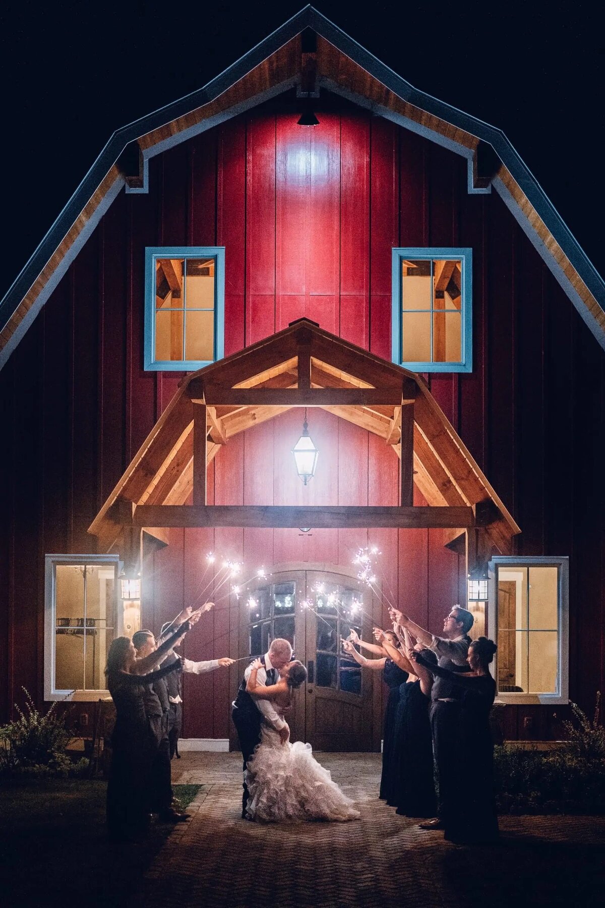 A rustic barn forms the backdrop for a magical wedding moment, with the bride and groom sharing a kiss as sparklers light up the night.