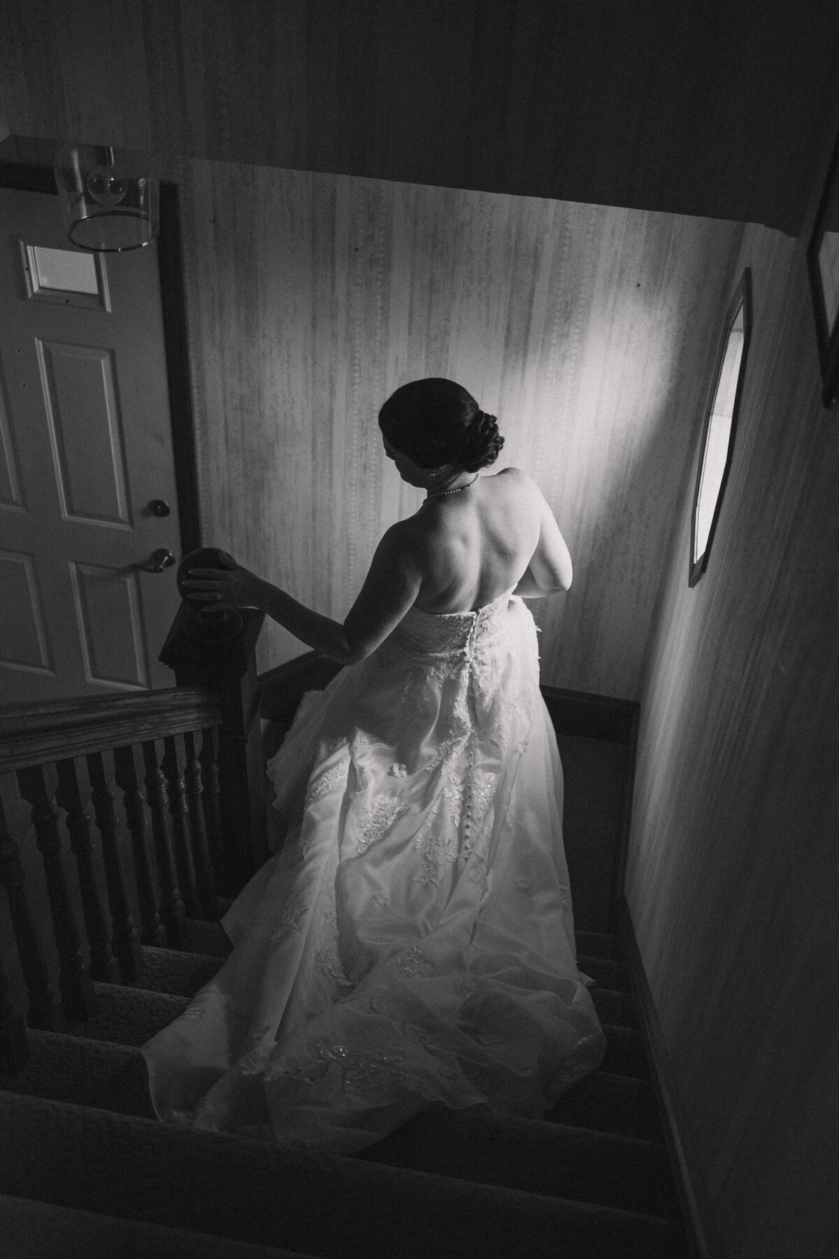Black and white image of a bride descending a staircase in her wedding dress, captured from behind to highlight the gown's intricate details and her elegant hairstyle