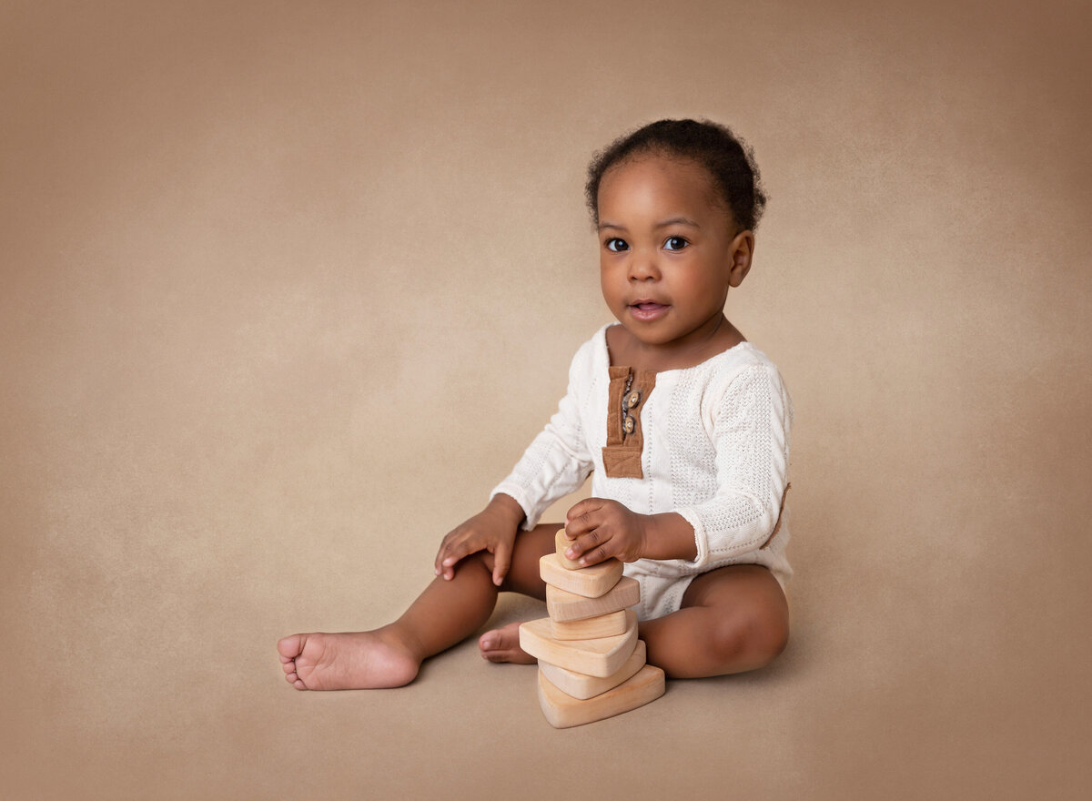 Baby is sitting for a 6-month milestone photoshoot. Baby is wearing a white knit long-sleeved romper with wooden toys stacked in front of him. Baby is looking at the camera with a happy expression.