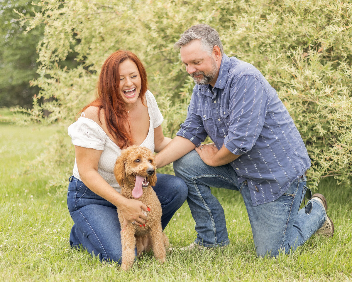 Mom and Dad are knelt down in grass in front of a bush with their light brown dog laughing and looking at the dog.  Dad is wearing blue jeans, blue and white stripped button up shirt, and has short pepper hair and beard.  Mom is wearing blue jeans, short sleeve white shirt, and has long red hair. 