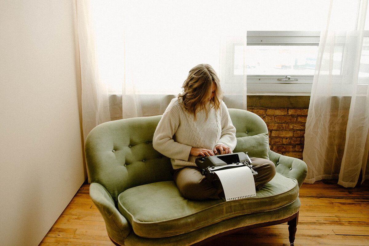 Brand designer sits on couch and types letter to client on her vintage typewriter.