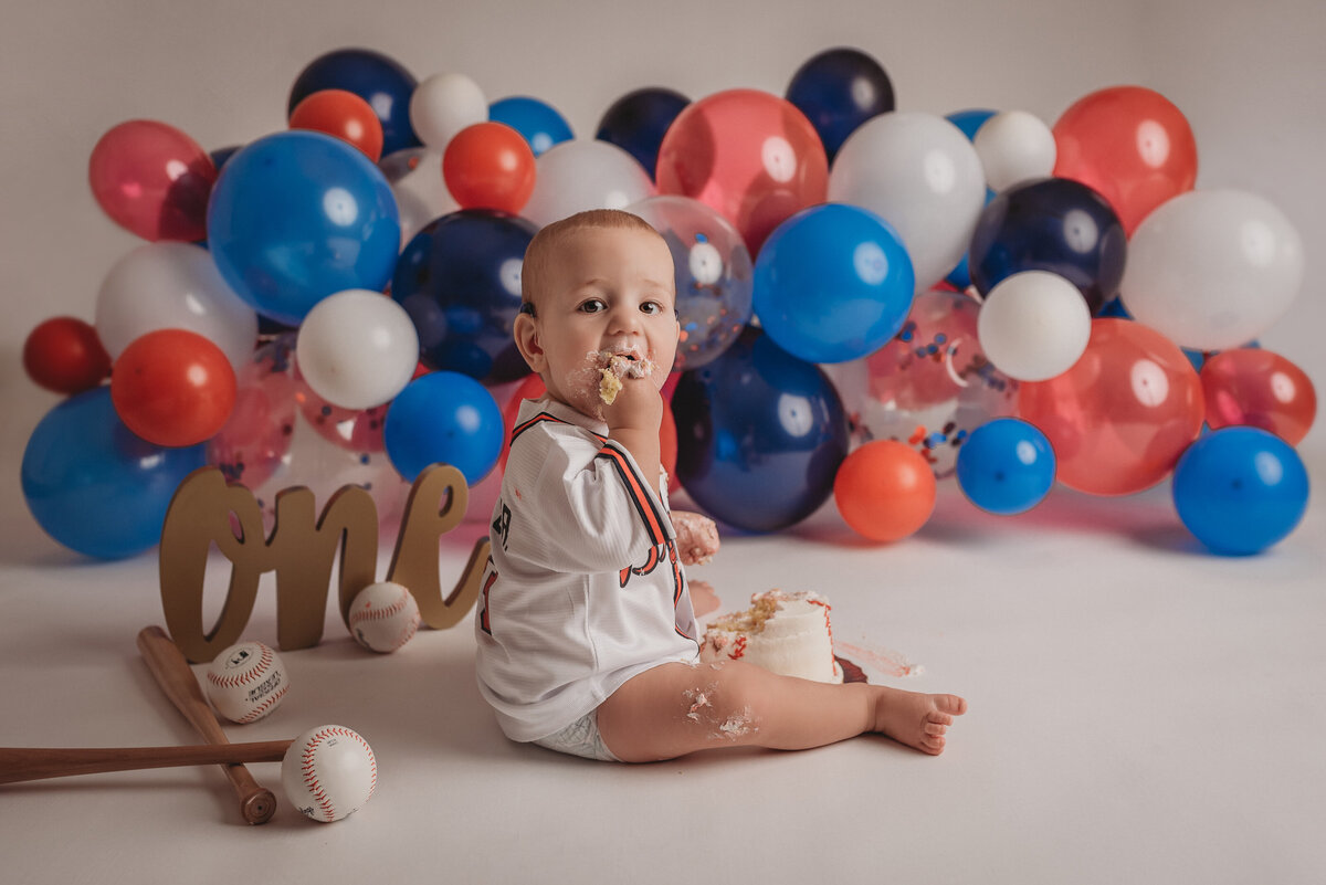 One year old baby boy sitting on floor eating birthday cake with Atlanta Braves baseball jersey on and red, white, blue balloon garland behind him