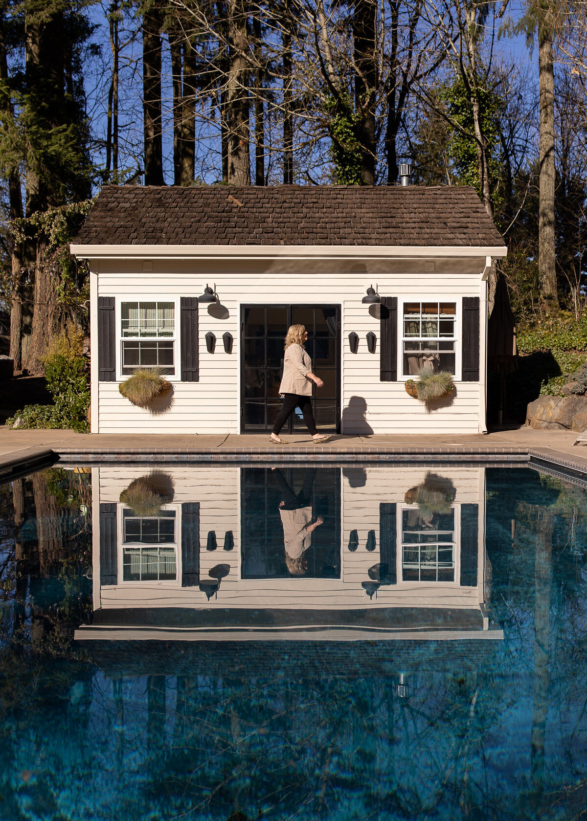 Woman walks passed pool house with her reflection in blue pool below her. 