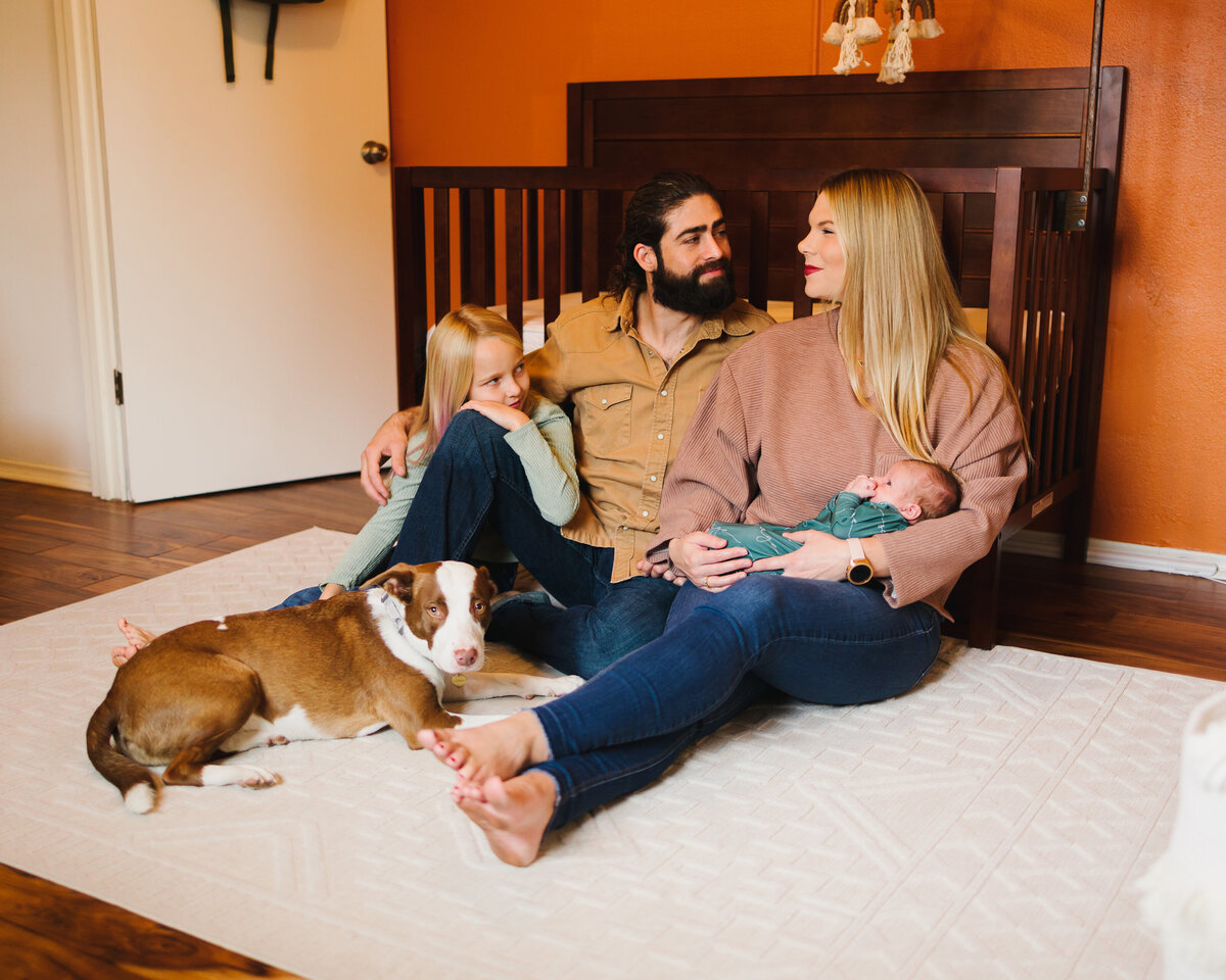 Family photo in a room recharged in the crib. The people are sitting on the floor, the woman is holding the newborn baby and is wearing a pink jacket and jeans. The man is looking at the woman and is wearing a yellow shirt. The girl on the left side is with her arm on the man's leg
