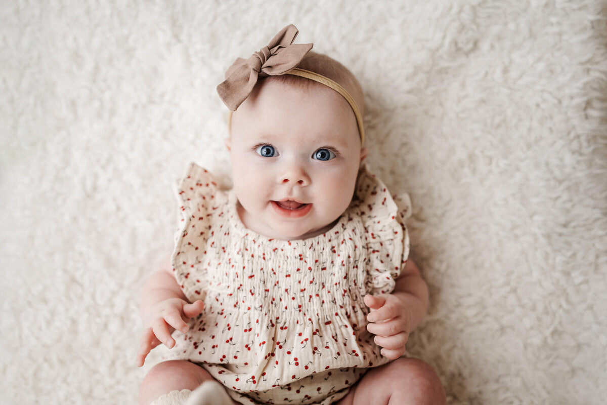 A baby in a light-colored outfit with red dots and a brown bow headband sits on a fluffy white surface, smiling and looking up with bright blue eyes. Captured by a talented portrait photographer in Bloomington, Illinois, this image is perfect for cherished families photography.