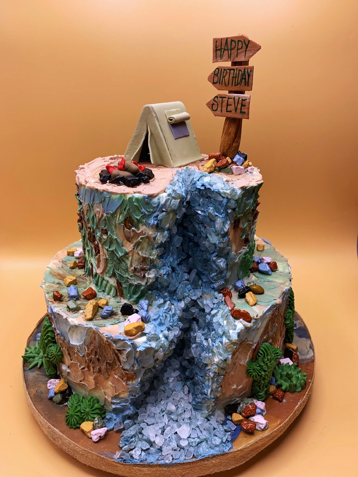 Outdoors hiking inspired 2 tier cake with  rock sugar waterfall, buttercream painted mountain background, succulents, with camping tent and campfire on top tier
