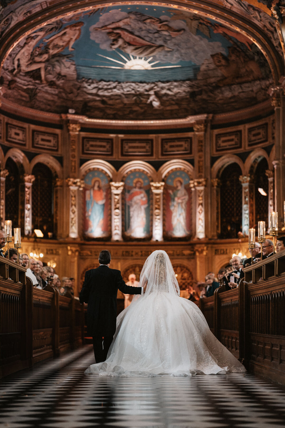 Bride and dad walk down the aisle at Royal Holywell Chapel in London, surrounded by guests in a richly decorated and historic chapel, showcasing an elegant and luxurious wedding celebration.