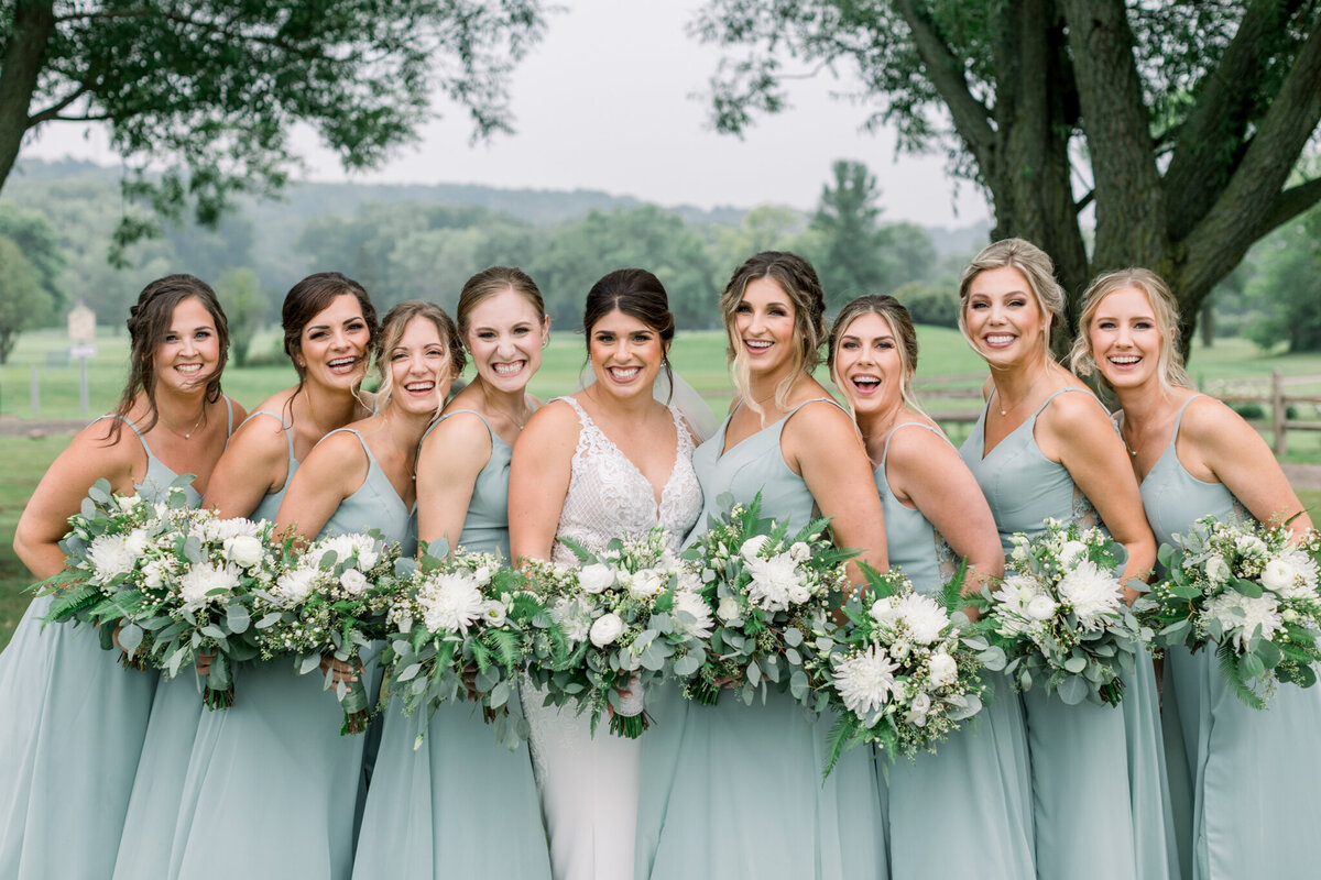 Bride in ivory (center) and eight bridesmaids in sage dresses holding white and green bouquets and laughing at the camera at a wedding in Green Bay, WI