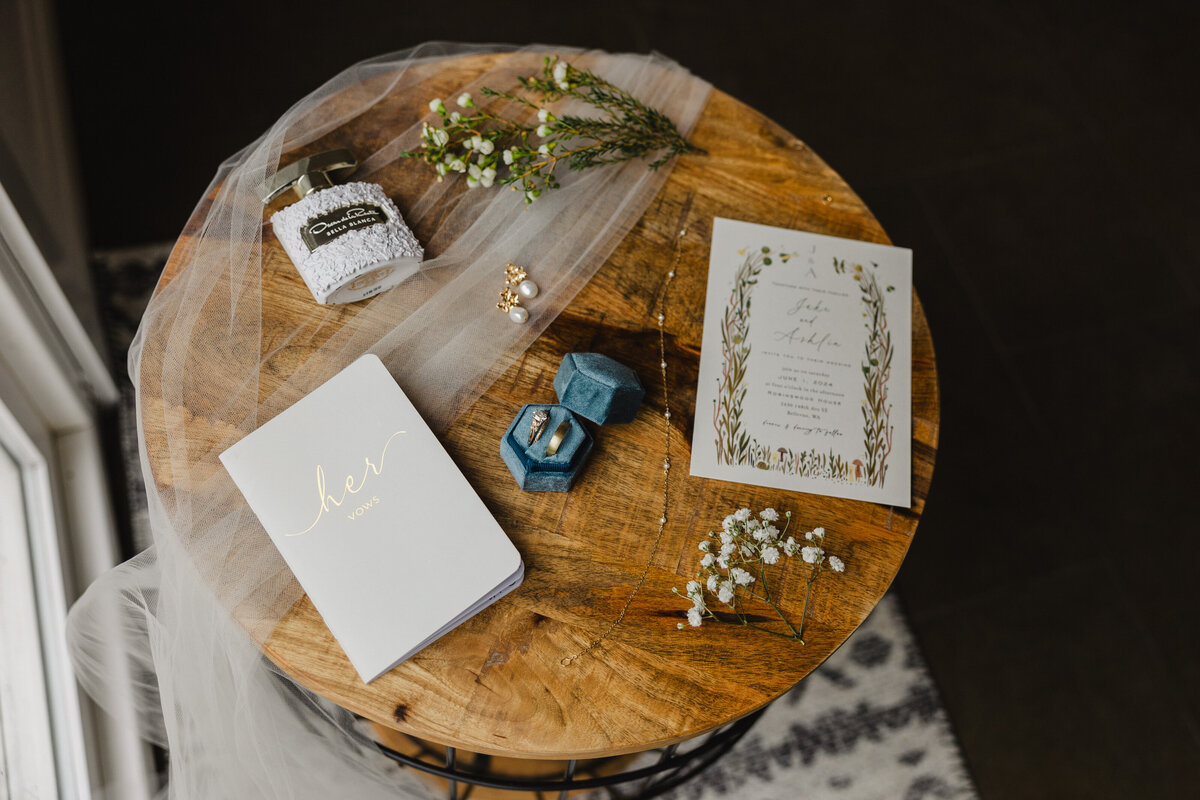 A wooden table with wedding invitations, a vow book, jewelry, wedding rings, perfume, a veil, and floral clippings