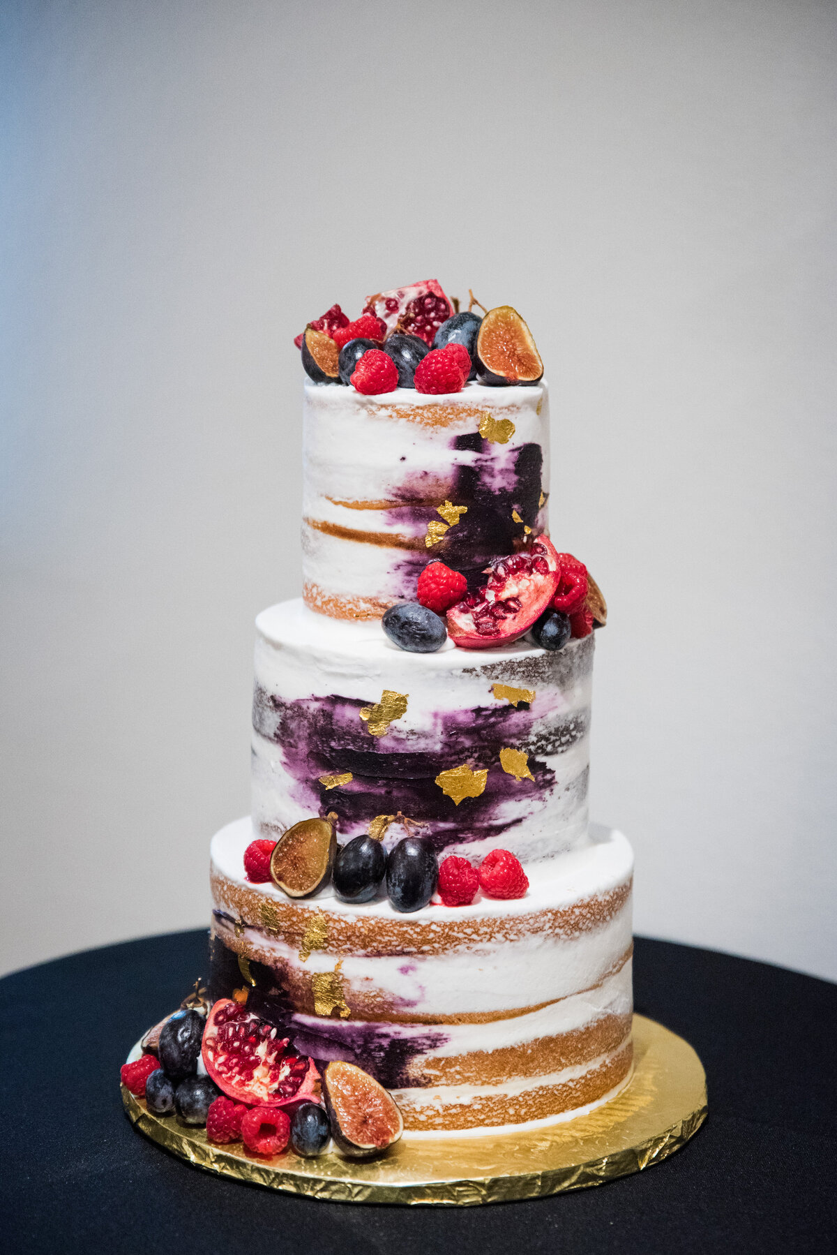 A three-tier wedding cake with partial white icing, raspberries, grapes, figs, and pomegranates.