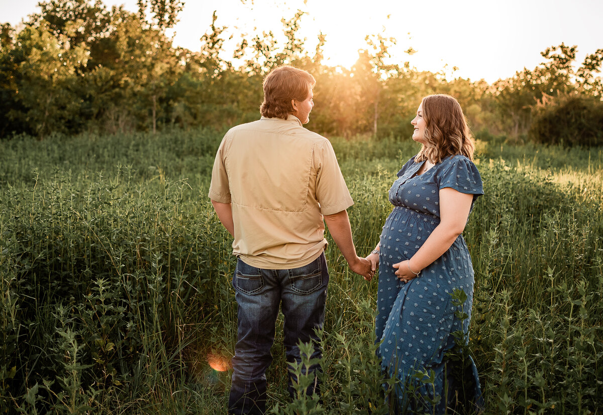 A pregnant wife looks at her husband as she cradles her belly in a field at sunset.