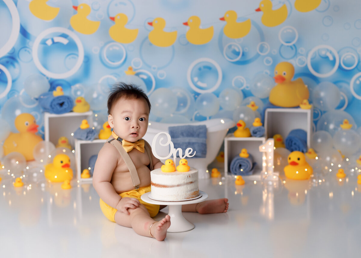 Rubber ducky cake smash in West Palm Beach and Boca Raton newborn and cake smash photography studio. Baby boy is looking at the camera sitting behind a white naked cake with rubber duckies. He is wearing a yellow diaper cover and bowtie with suspenders. The background is a bubble inspired drop with a small claw foot tub surrounded by rubber duckies and blue hand towels.