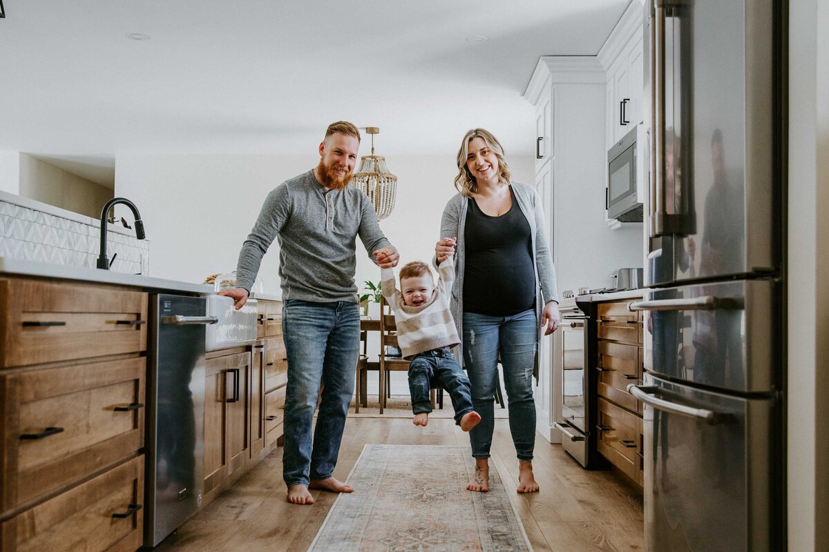 In home photo session - Expectant mom, dad, and toddler in their kitchen. Mom and dad are swinging their little boy between them. Boy has joyful smile.