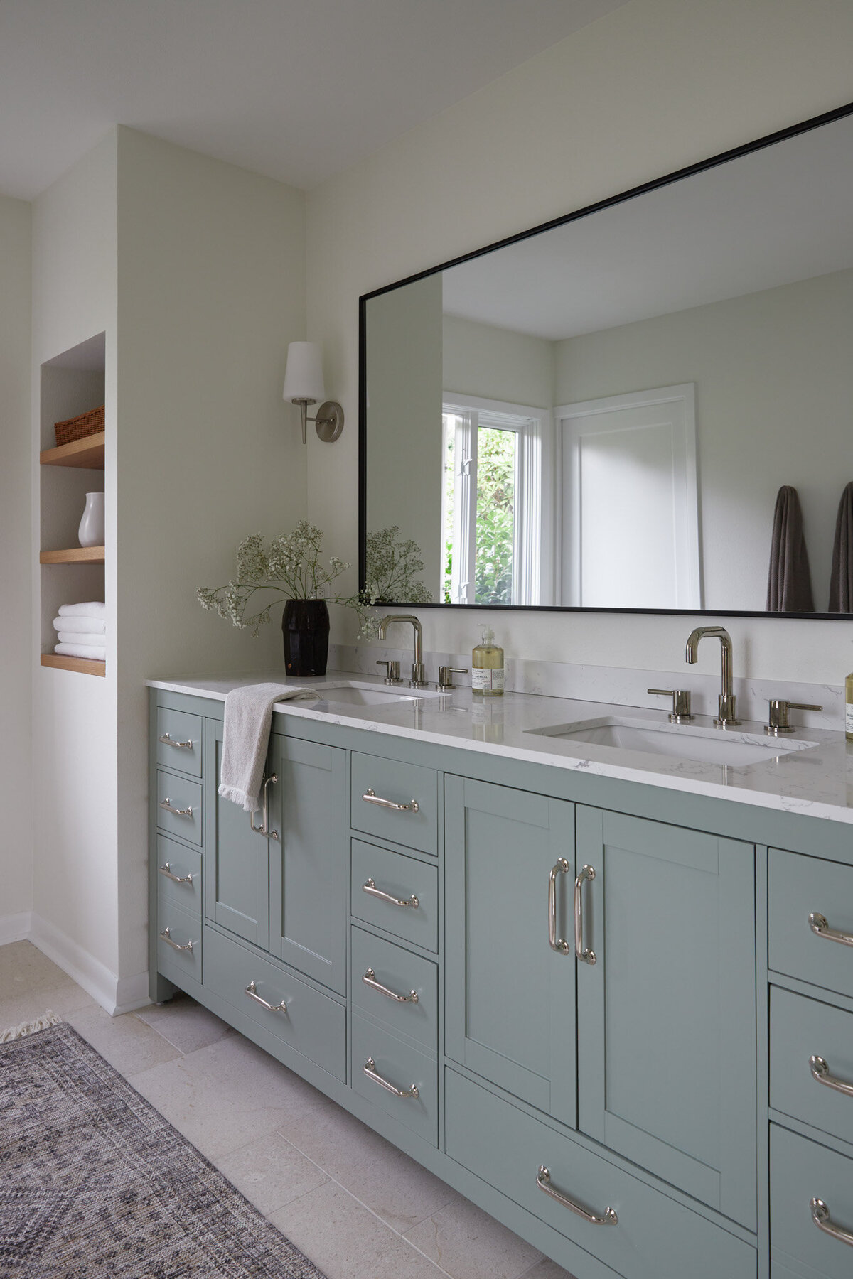Bathroom with light blue bathroom vanity with silver cabinet pulls. A silver sink faucet with a black mirror above. Next to the sink is a dark brown vase with baby’s breath flowers, a Handsoap pump, and a white candle. Next to the vanity is a white wall with recessed white oak shelves with photos, jars, and a woven basket.