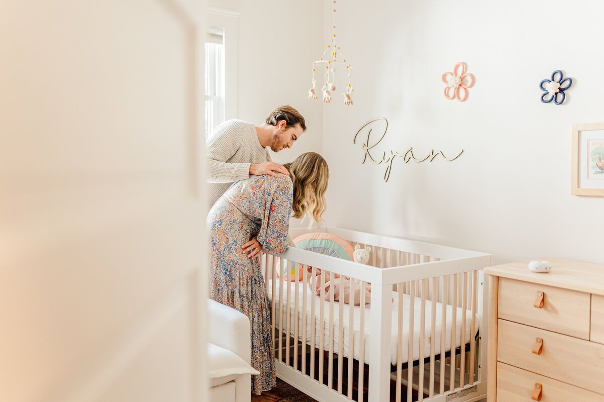 Seen through a doorway, two parents lean over a crib and smile at their daughter in a session by a Boston Newborn Photographer