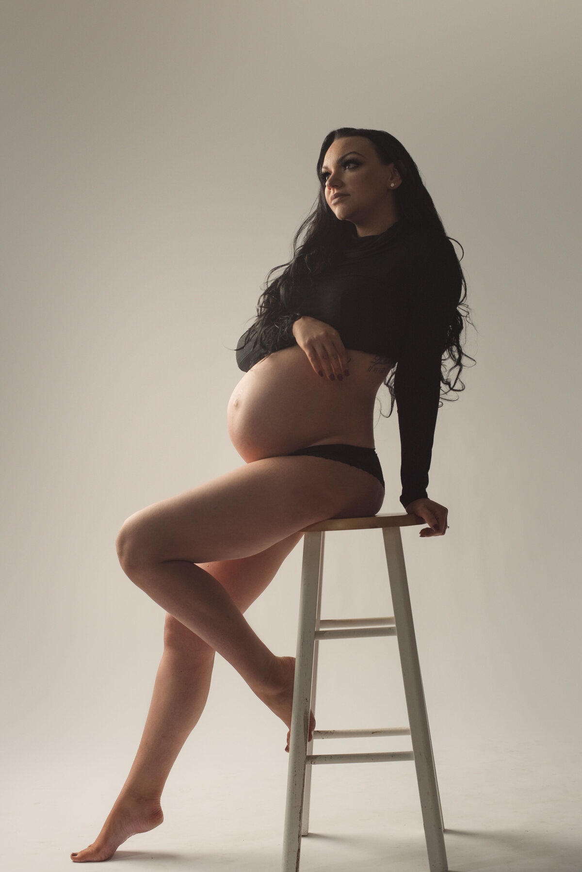 Pregnant lady at 34 weeks sitting on stool posing with one foot on stool holding baby bump and wearing long sleeve black crop top and black panties